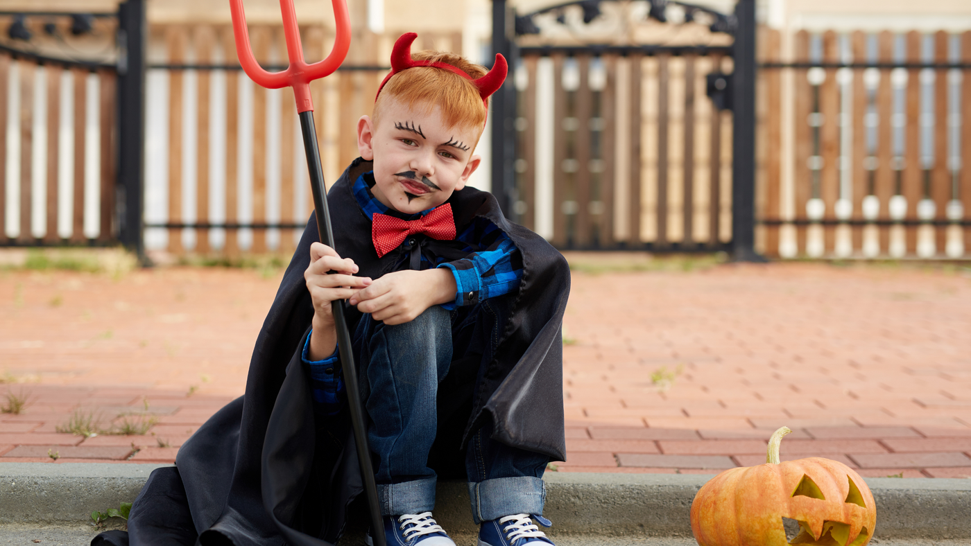 little boy sitting on kerb beside pumpkin dressed as a devil for halloween