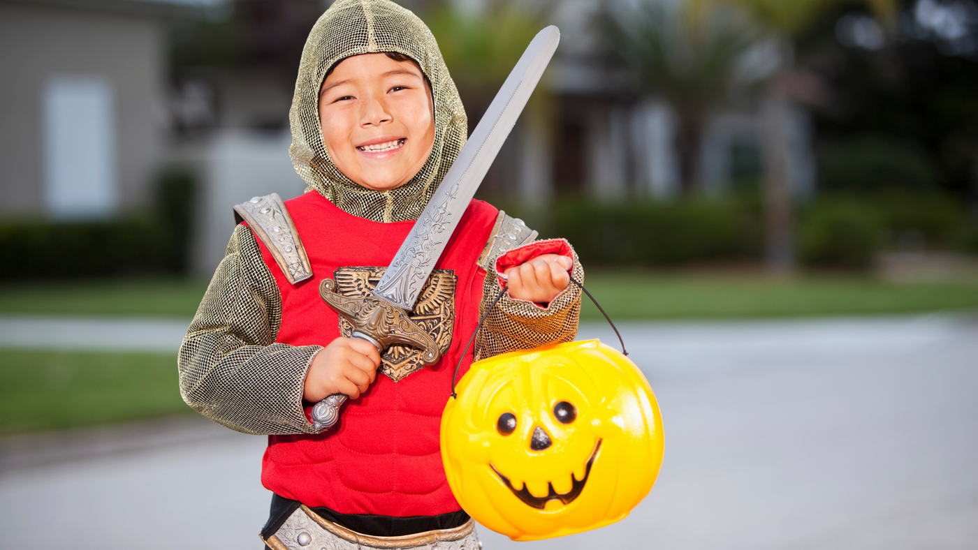 little boy dressed as a medieval knight on halloween holding an artificial sword and a pumpkin candy bag