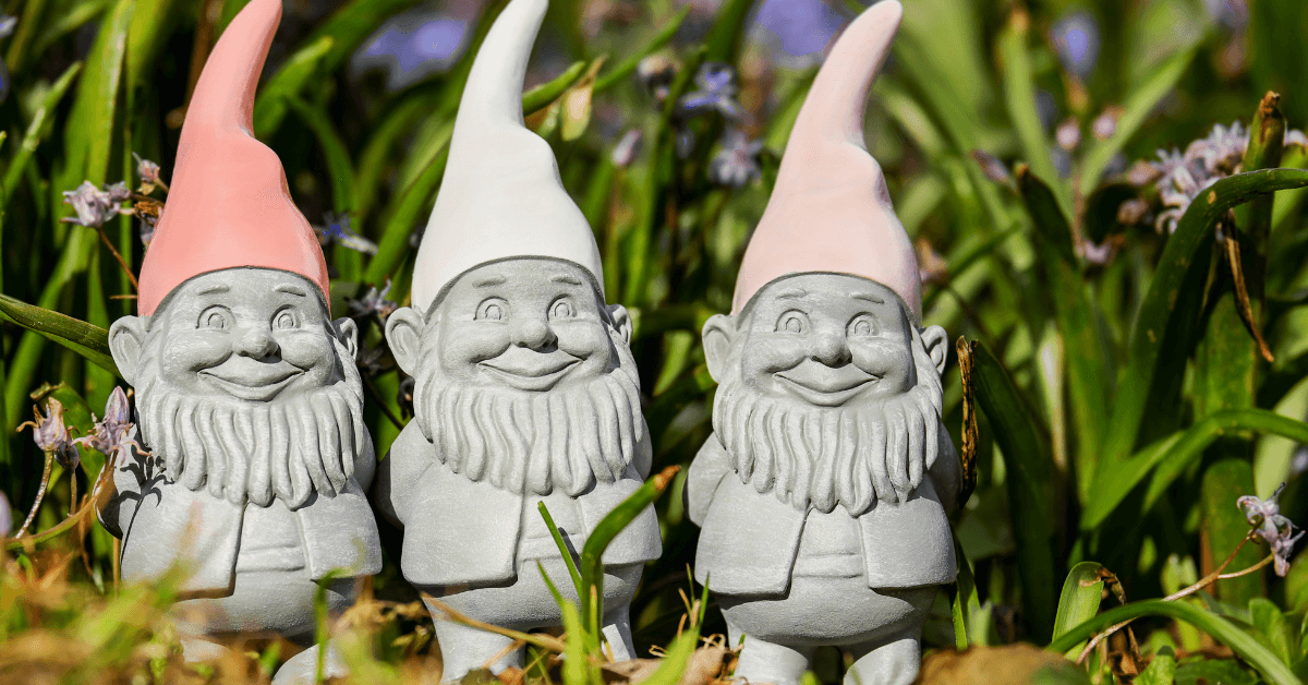 three garden gnomes in clay colour wearing pink and white hats resting in garden foliage