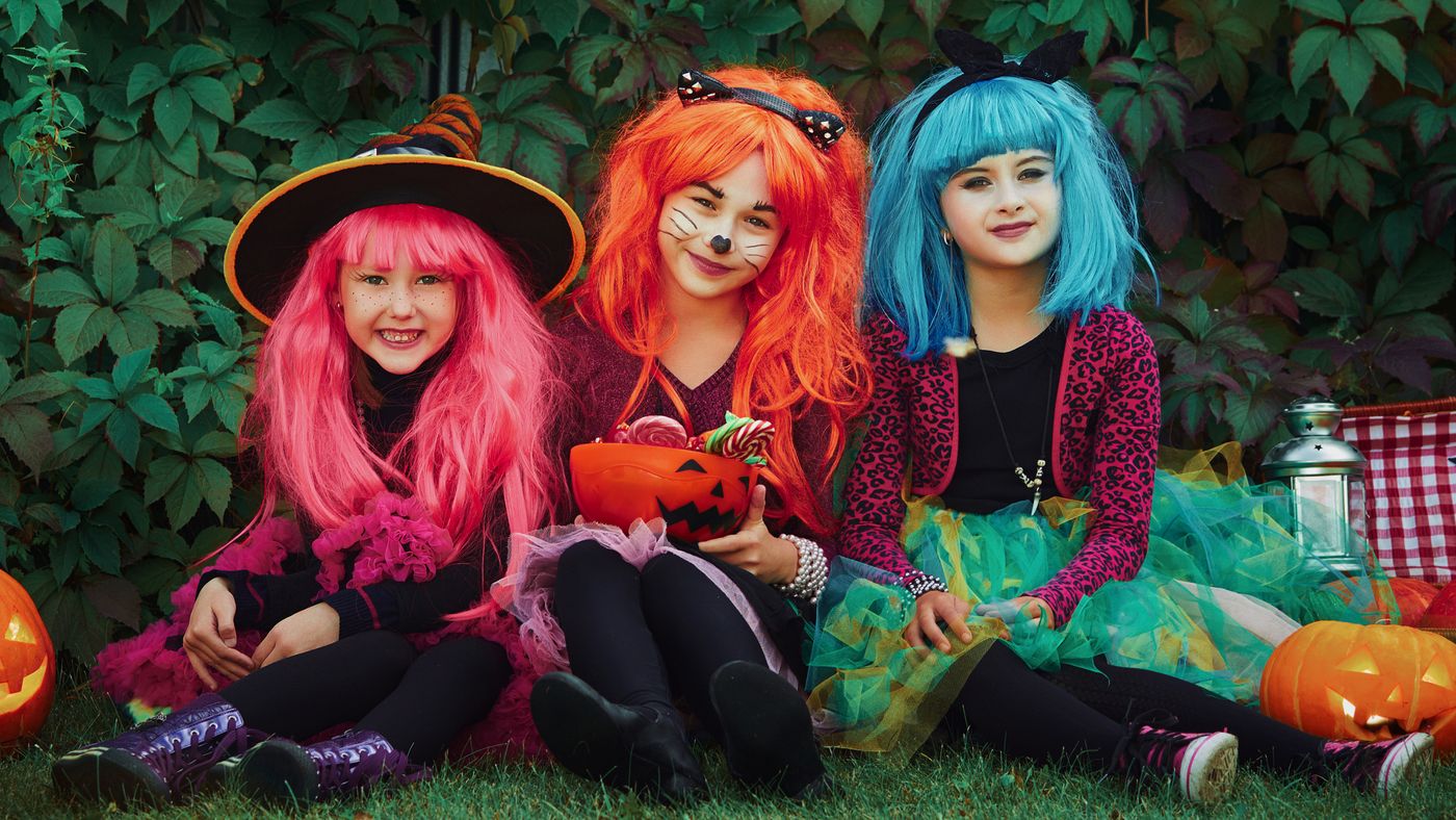 three little girls dressed for halloween in the garden wearing bold colour wigs in pink, orange and blue