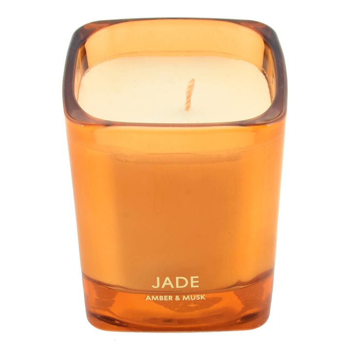 Orange candle in a glass holder labeled 'Jade Amber & Musk' on a white background