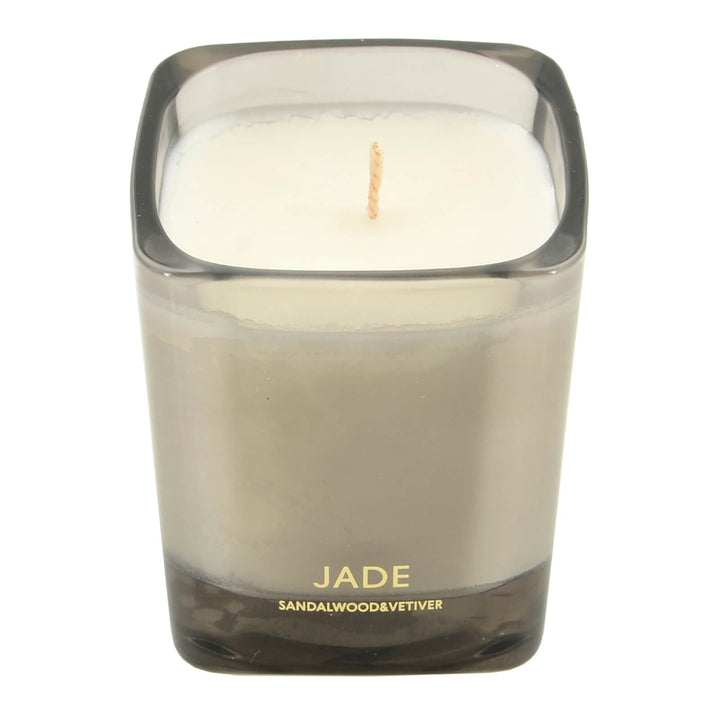 Candle in a grey glass holder labeled 'Jade' with sandalwood and vetiver scent on a white background