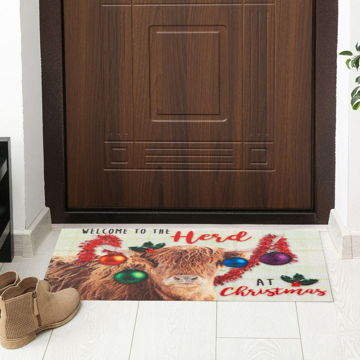 Doormat with a festive design of a highland cow wearing Christmas ornaments in front of a wooden door.
