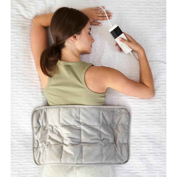 Woman lying on a bed with a grey heat pad on her back and a controller in hand