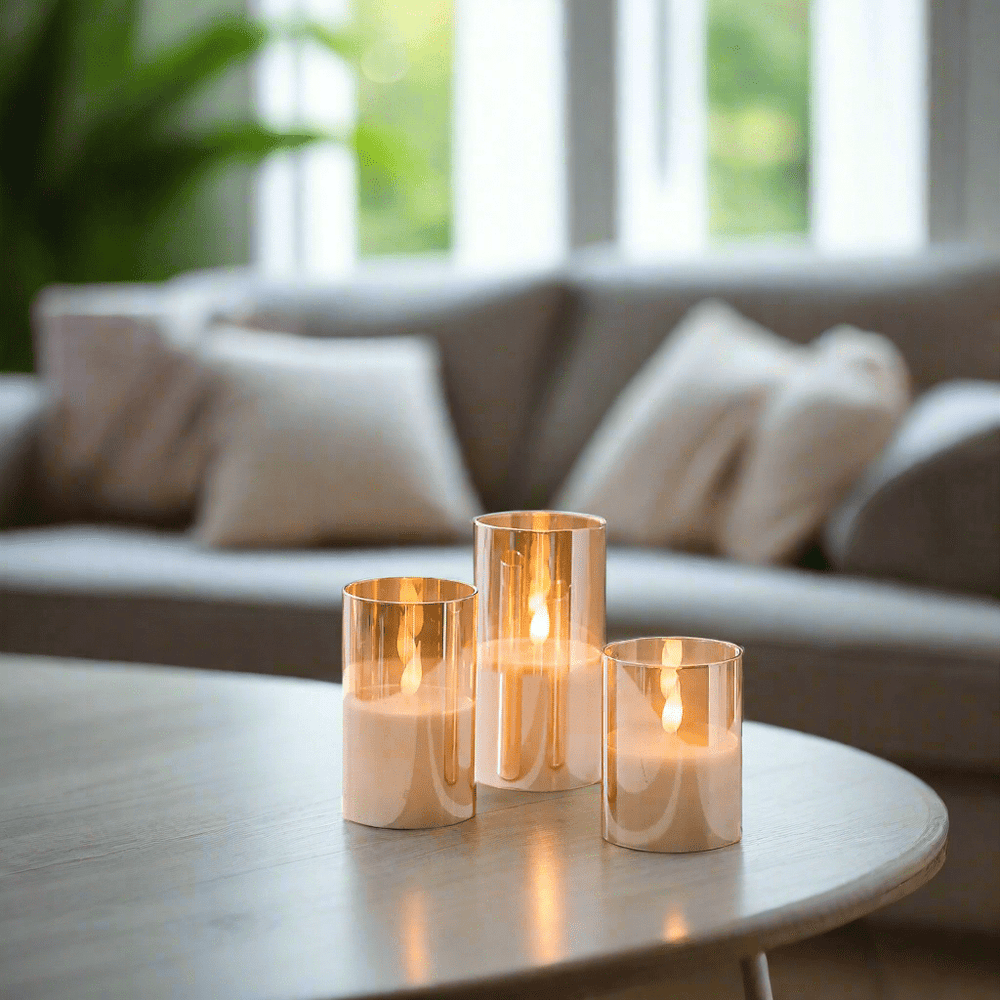 3 gold flameless candles on a wooden table in front of a sofa in living room with window visible in background