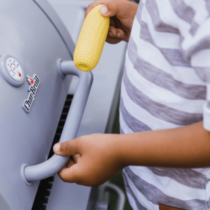 close up shot of little boys hand holding pretend corn on the cob in one hand, with other hand on BBQ handle at the front