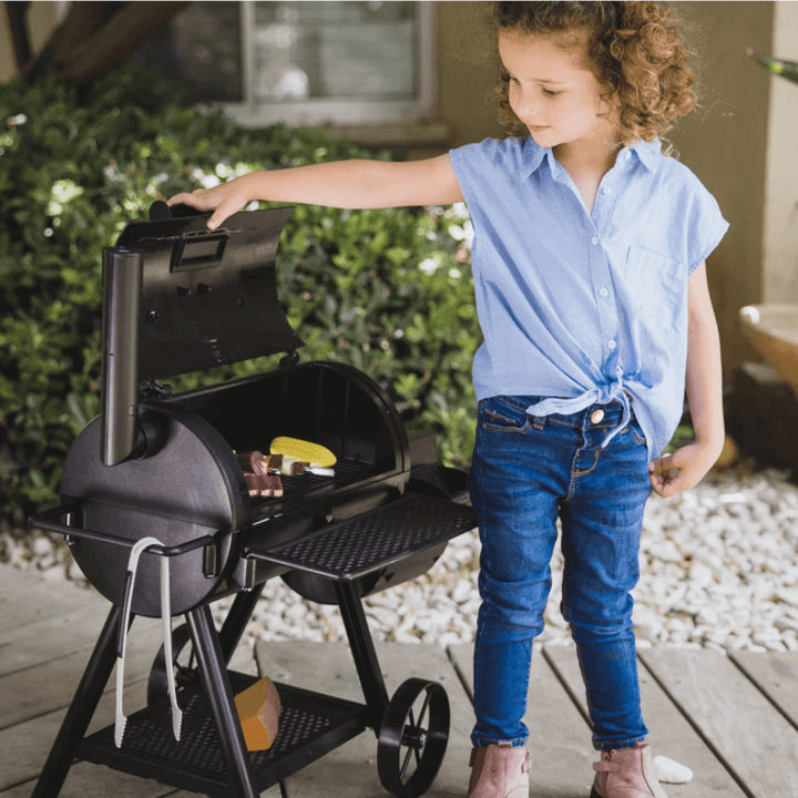 young girl in garden lifting lid on pretend smoker bbq to reveal pretend food on the inside