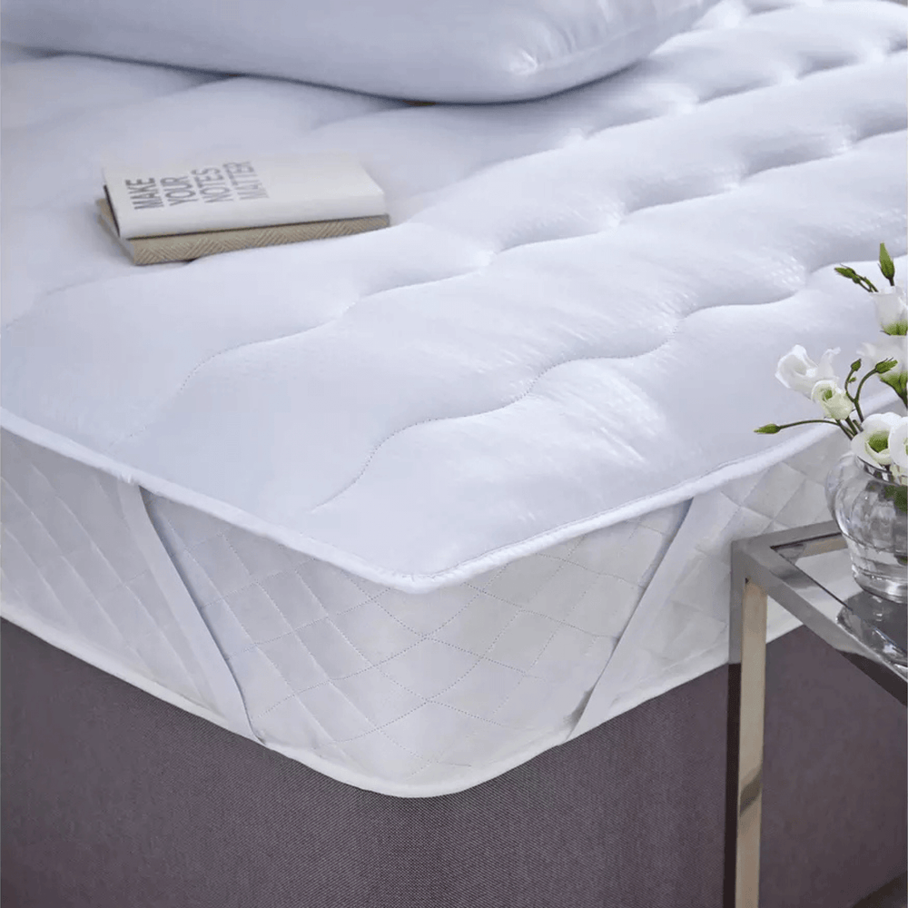 White quilted mattress protector on a bed with a small table and flowers in the foreground.