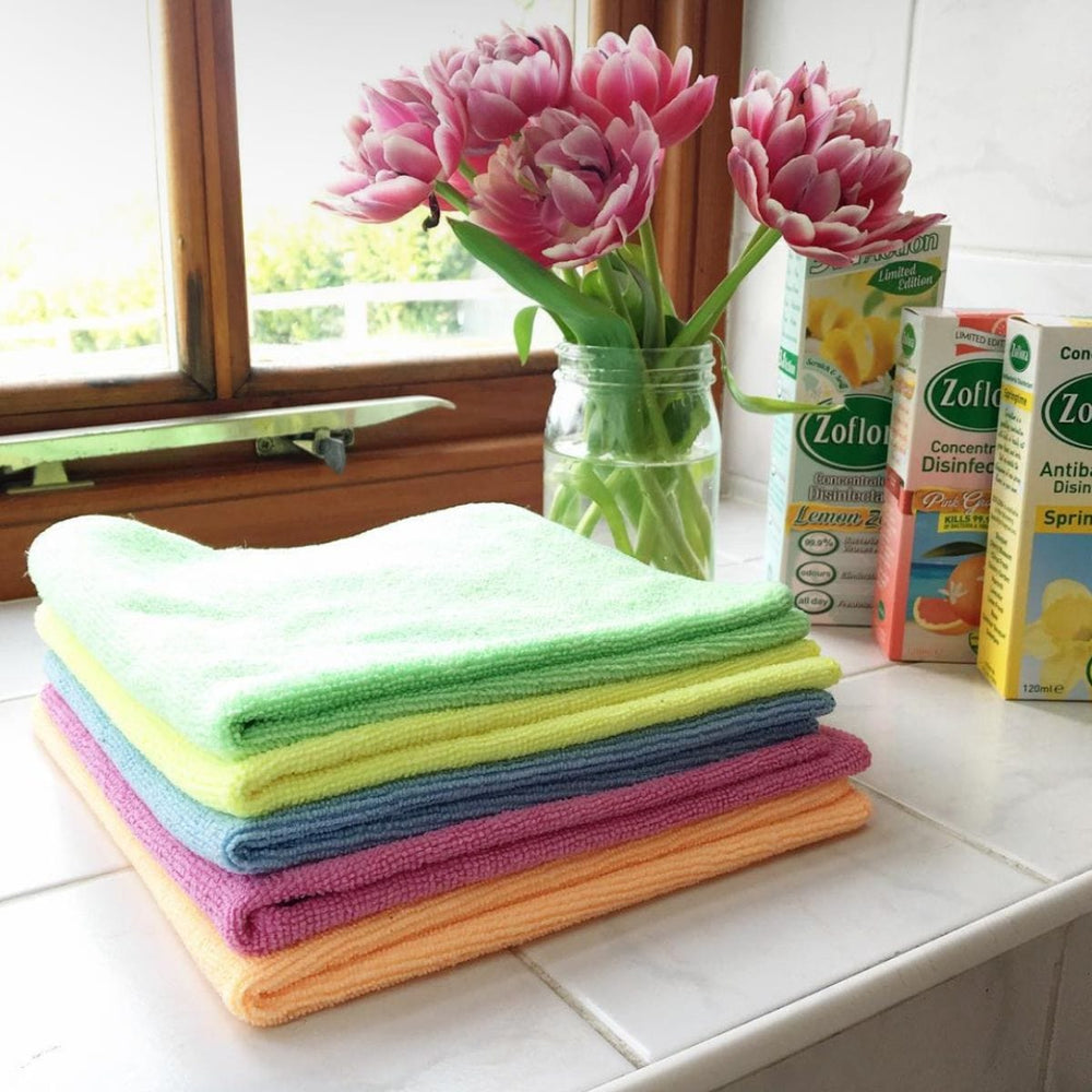 pile of cleaning cloths in green, yellow, blue, pink, orange colours on window ledge beside pink spring flowers and Zoflora cleaning products