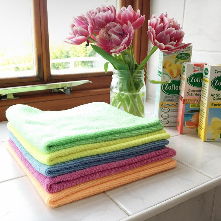 pile of cleaning cloths in green, yellow, blue, pink, orange colours on window ledge beside pink spring flowers and Zoflora cleaning products