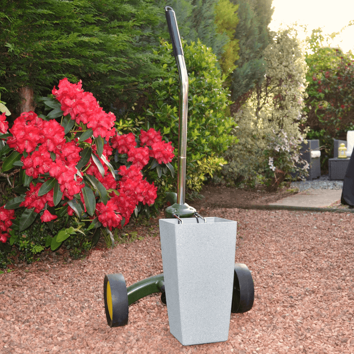plant pot mover with grey plant pit secured on red garden stones with red flower bushes and trees in background, rattan furniture visible further back