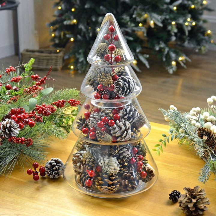 christmas tree storage jar on wooden coffee table with tree in background, filled with pine cones and red berry clusters