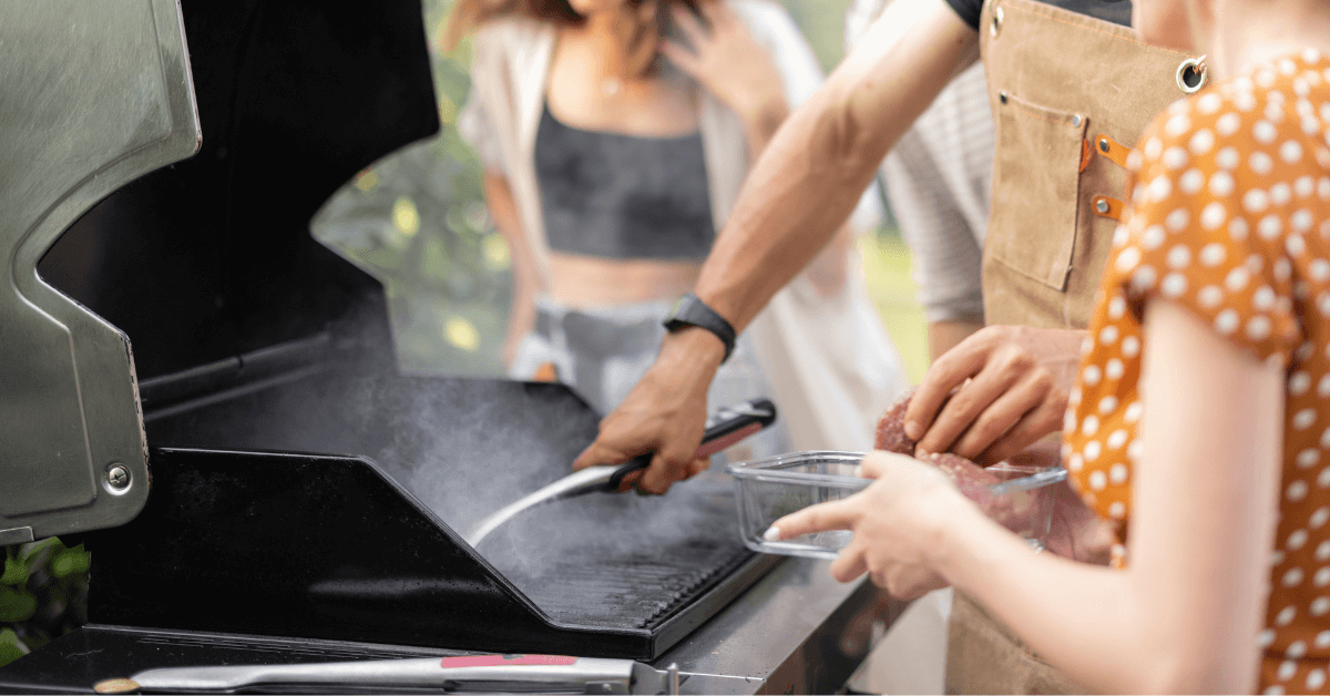 unidentified people standing in front of charcoal bbq cooking meat with bbq tongs resting on the side
