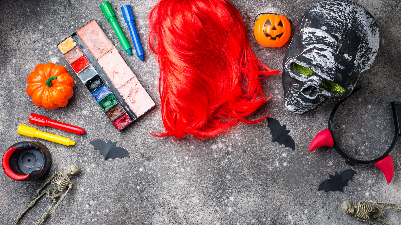 halloween accessories including red wig, devil horns face paints and mini pumpkins on a grey background