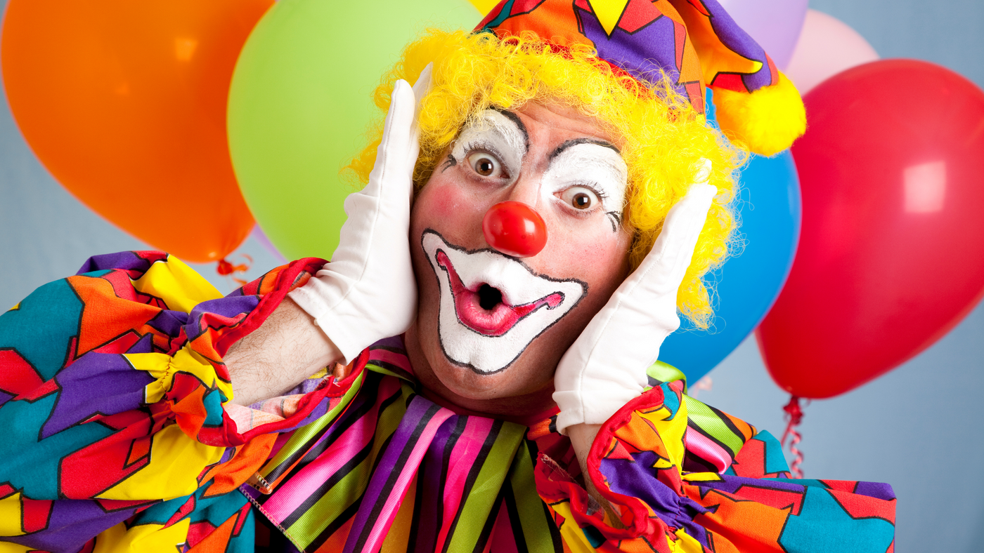 circus clown wearing bold colours and standing in front of multi coloured balloons