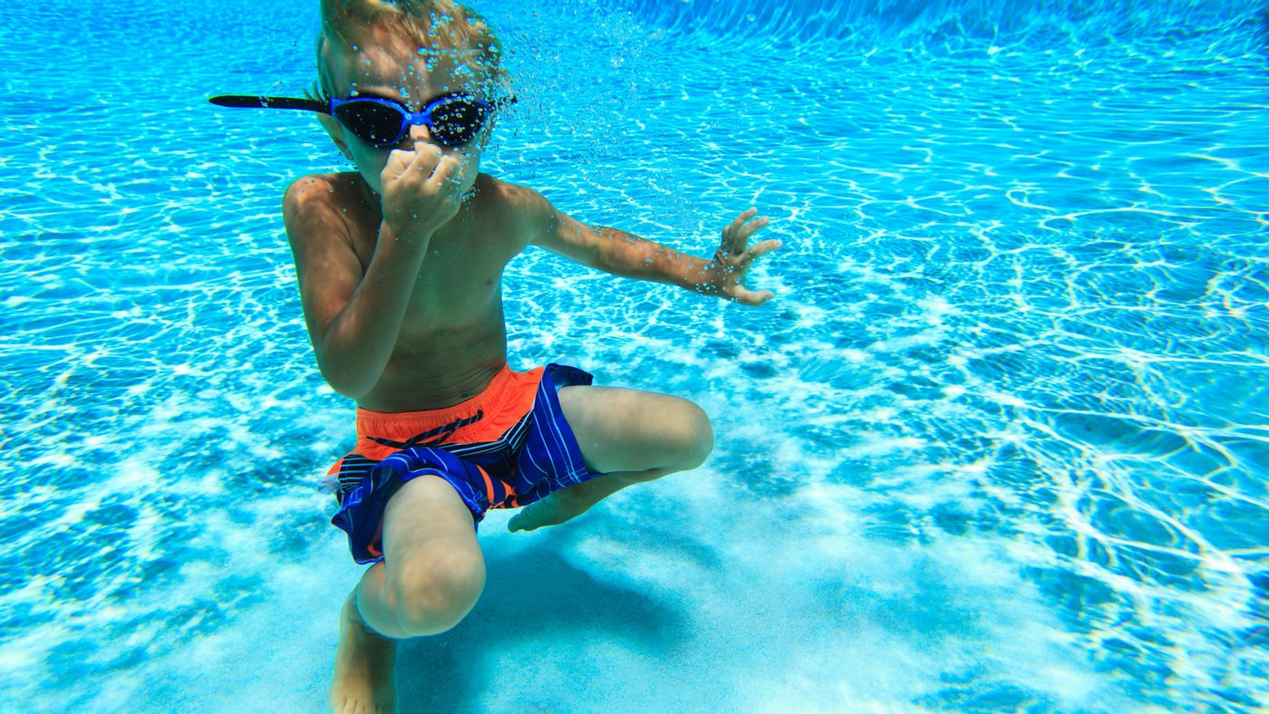 little boy in pool water wearing swim shorts and swimming goggles