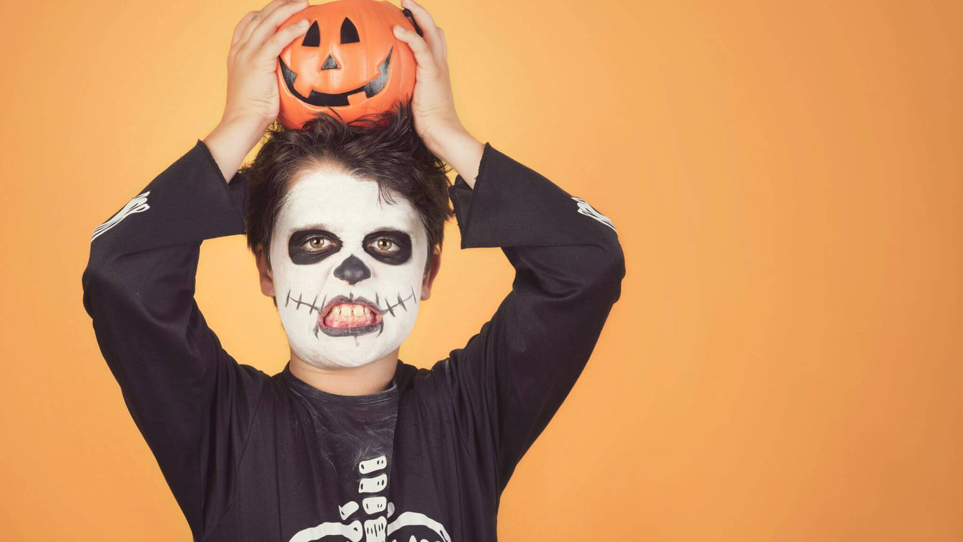 young boy dressed as a skeleton with black and white face paint holding a PVC pumpkin on his head, shot on an orange background