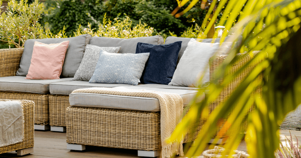 brown coloured rattan sofa in garden space featuring colourful cushions and palm leaves in foreground
