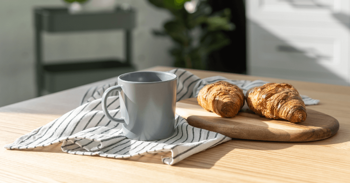 grey coffee mug on wooden kitchen counter beside chopping board featuring 2 breakfast croissants