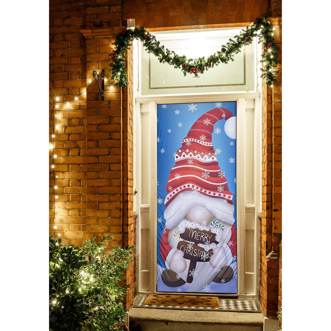 Decorative Christmas door cover with a gnome holding a 'Merry Christmas' sign, lit by string lights on a brick wall.