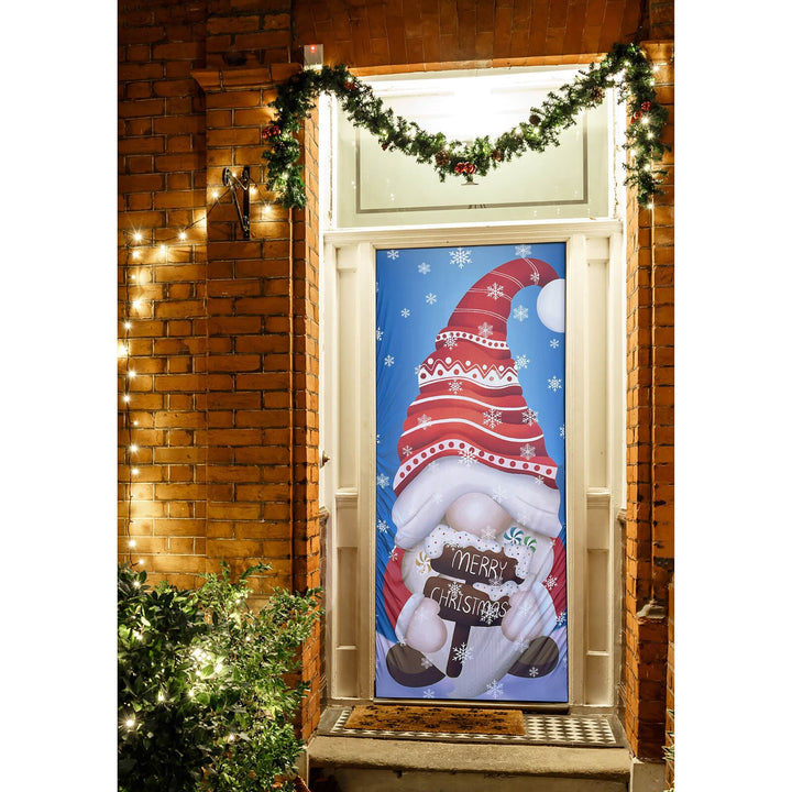 Decorative Christmas door cover with a gnome holding a 'Merry Christmas' sign, lit by string lights on a brick wall.
