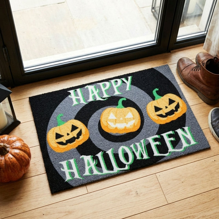 A rectangular Halloween doormat placed on a light wood floor in front of a glass door. The mat is black and gray with the words “HAPPY HALLOWEEN” in white and green, plus three orange jack-o'-lantern pumpkins.