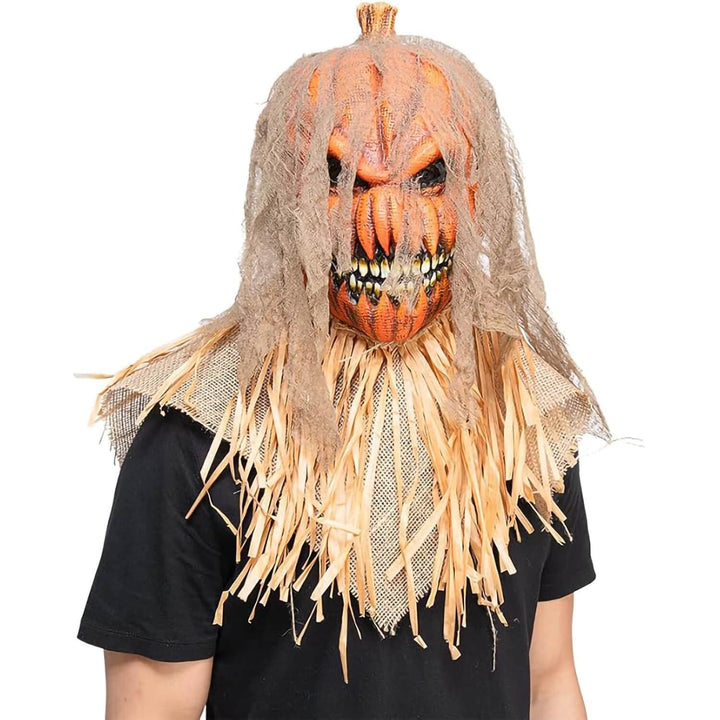 A scary Halloween scarecrow mask with a carved orange pumpkin head, tattered beige netting, and straw-colored hair, shown worn by a person against a plain white background.