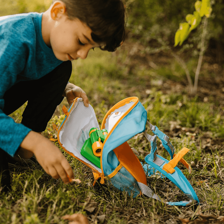 young boy in outdoor location packs his pretend play set into his blue and orange hiking backpack