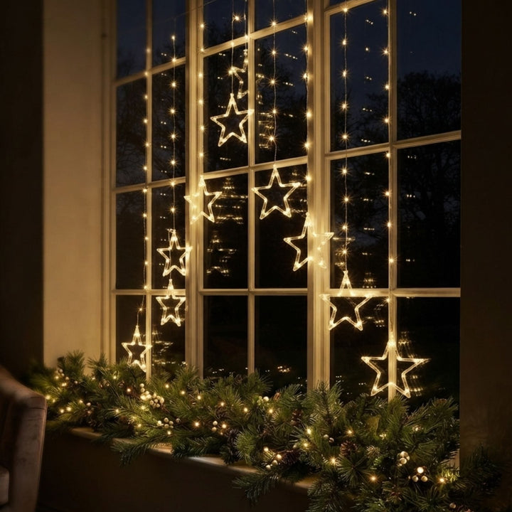 Warm white LED star string lights hanging on a large window, with a green garland of similar lights draped along the window sill.