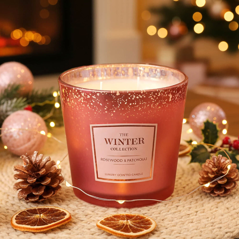 A Rosewood & patchouli scented rose gold colour metallic candle jar with a glittery finish and on a table with dried orange slices, pinecones, fairy lights, and holiday decorations in the background.