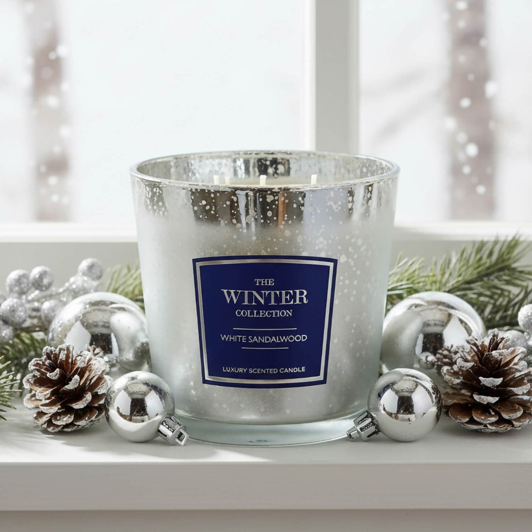A white sandalwood scented candle in a silver glittery glass jar, styled on a white windowsill with silver ornaments, pinecones, and green pine branches. 