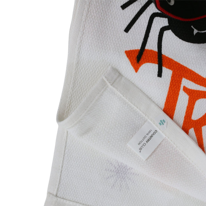 Close-up of a white textured fabric garment with a black spider graphic, orange lettering, and a sewn-in size tag reading '100% cotton'.