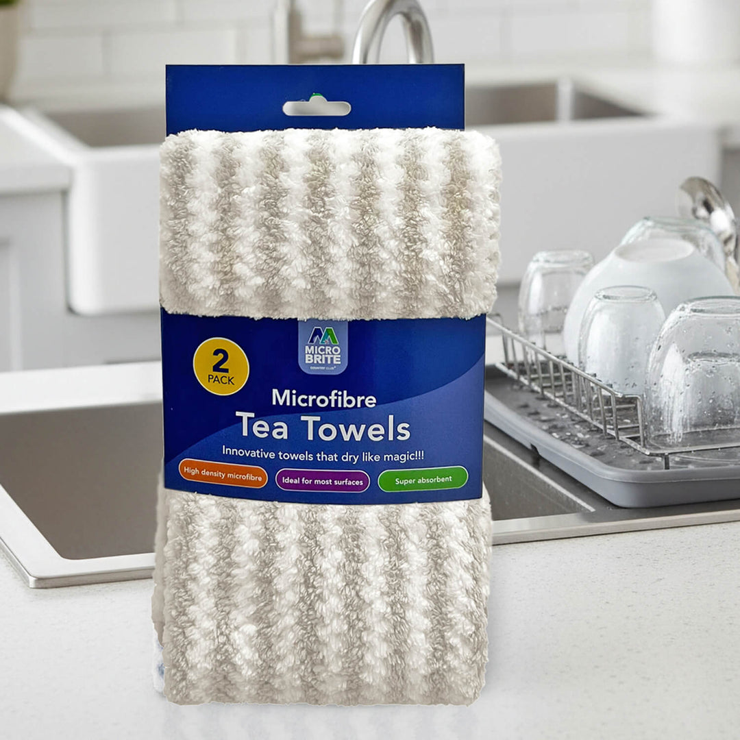 A 2-pack of Micro Fibre Tea Towels in a cream/off-white textured fabric, packaged in a blue cardboard sleeve with a sink and drying rack in the background.