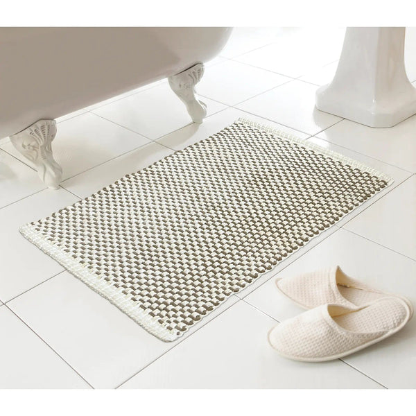 Beige and white woven style bath mat on a white tiles floor beside slippers and a bath