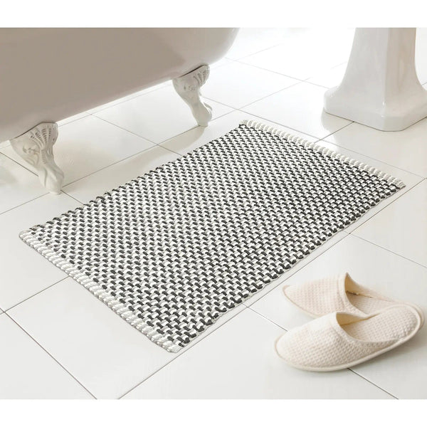 Dark grey and white woven style bath mat on a white tiles floor beside slippers and a bath