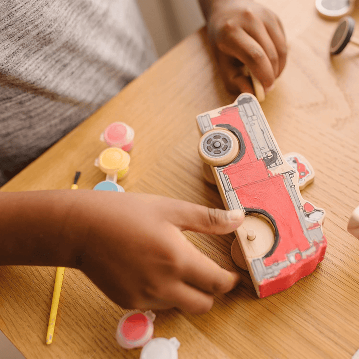 young boy adds wheels to wooden block to create fire engine craft