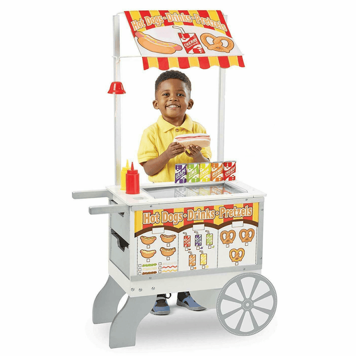 little boy stands behind hot dog stand holding a fabric hot dog with wooden soda cans beside him