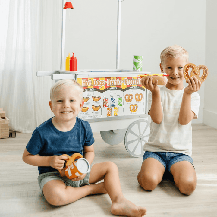 two little boys smile sitting in front of hot dog cart holding pretend wooden pretzel fabric hot dog and wooden soda can