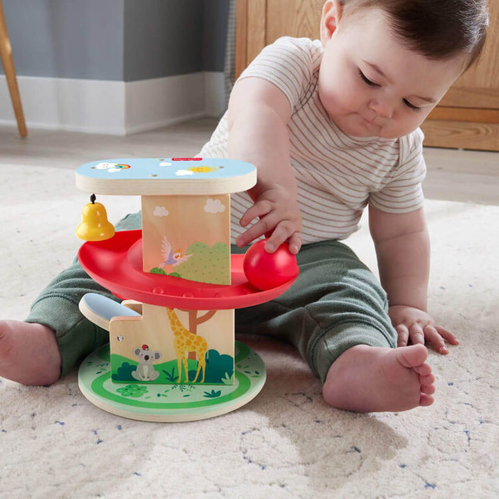 Baby playing with a wooden treehouse playset with red ball and yellow bell toy on a carpeted floor.