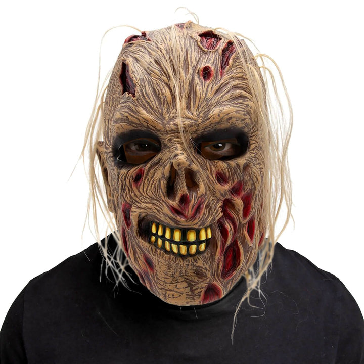 A horror zombie mask with tattered beige skin, bloody wounds, and a yellowed tooth grin, worn by a person in a black shirt against a plain white background.