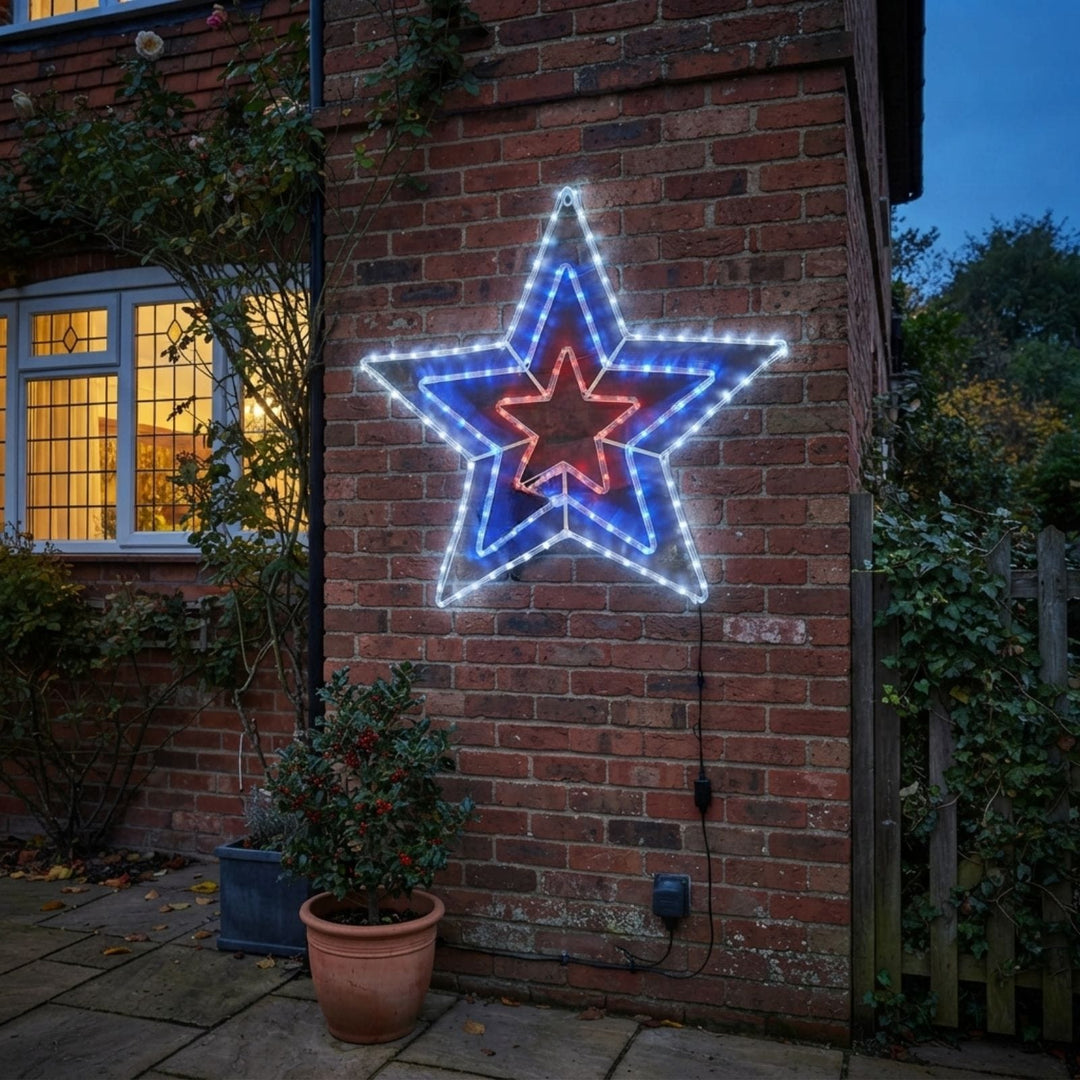 A large five-pointed star-shaped LED light decoration mounted on a red brick wall at dusk, with white outer rings, a blue middle ring, and a red center ring. A lit window, potted plants, and garden foliage are visible around it.