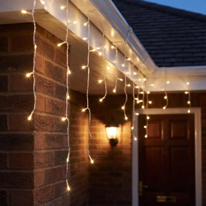 Warm white icicle string lights hanging from a brick house exterior at dusk, glowing softly near a door and wall sconce.