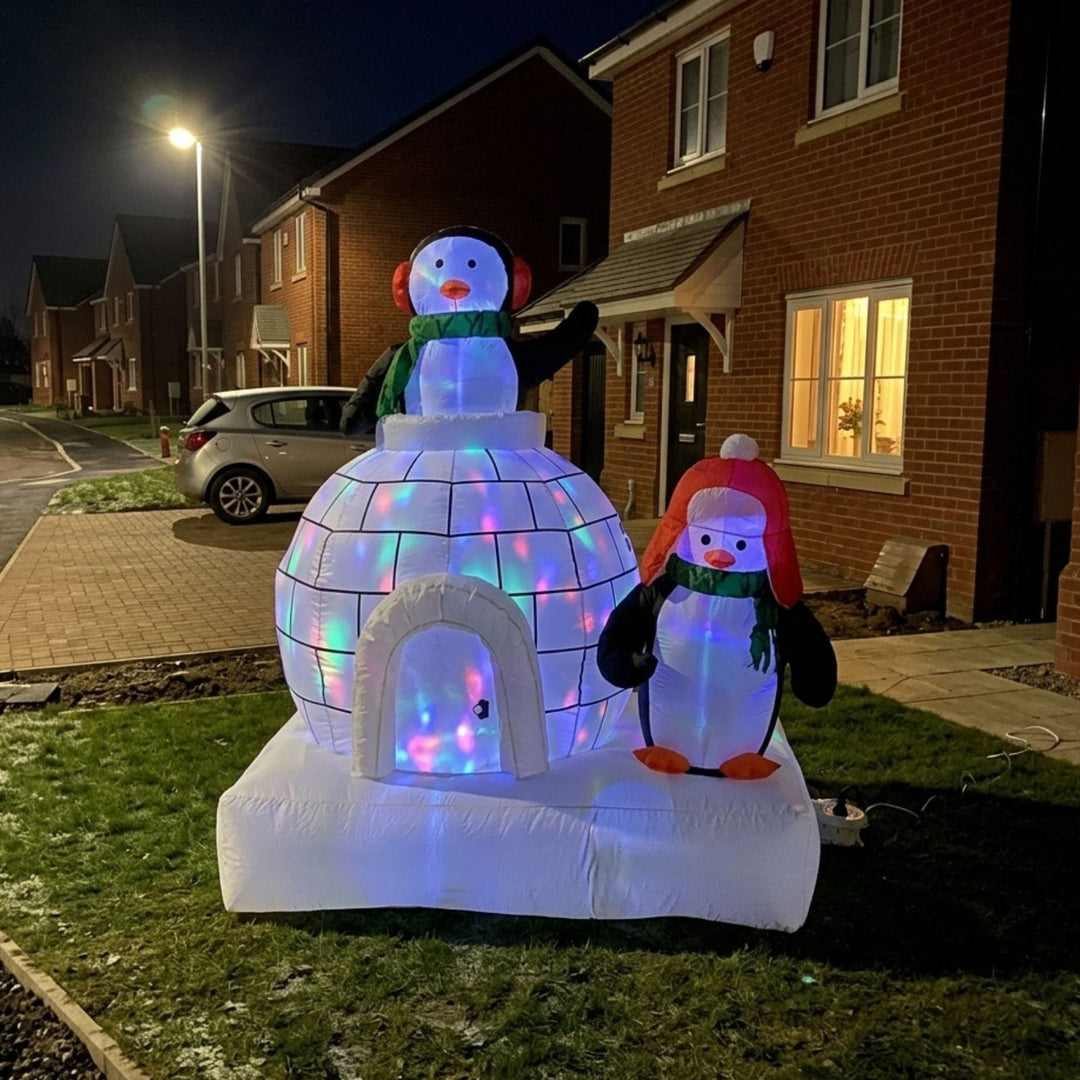 An inflatable Christmas decoration set featuring a glowing white igloo with a snowman and a penguin, photographed outdoors on a lawn in front of residential houses at night.