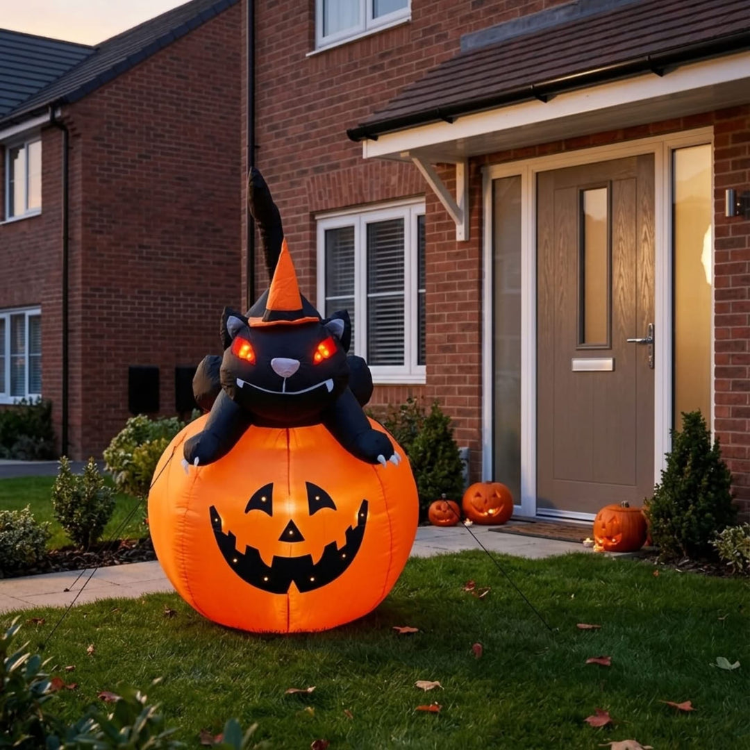 An inflatable Halloween decoration featuring a black cat wearing an orange witch hat sitting on a large orange jack-o'-lantern, displayed on a lawn in front of a brick house.
