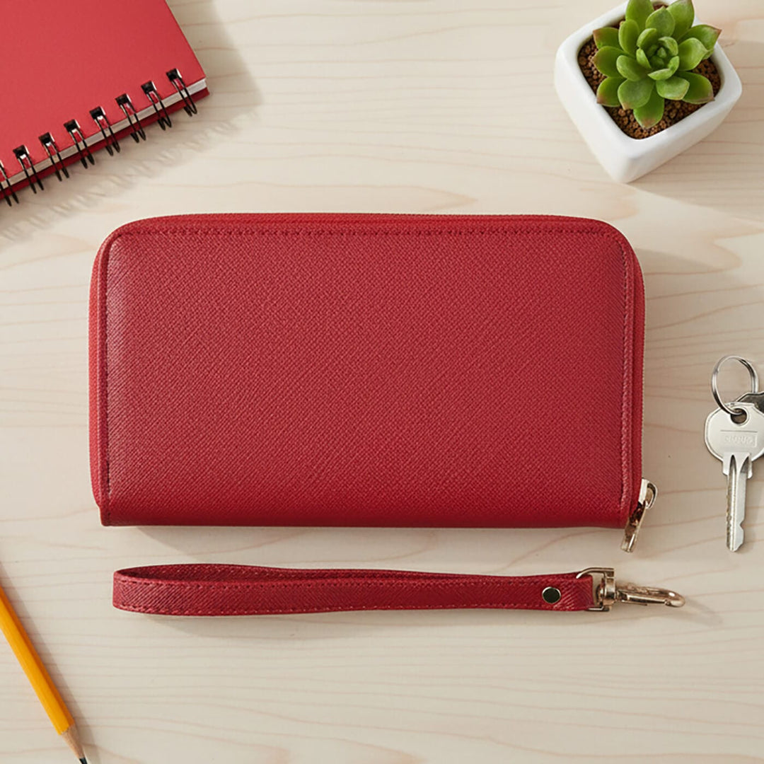 Red leather purse with wrist strap on a wooden surface with a notebook, plant, and keys.