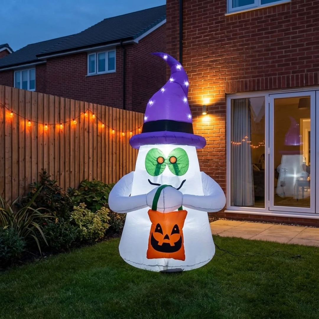 A large inflatable Halloween ghost decoration stands on a lawn in front of a brick house at dusk. The ghost is white with a purple witch hat, green oval glasses, a black mustache, and holds an orange pumpkin bag.