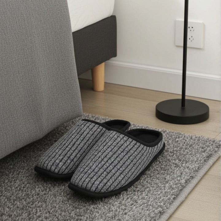 Pair of gray slippers on a rug next to a bed in a bedroom setting.
