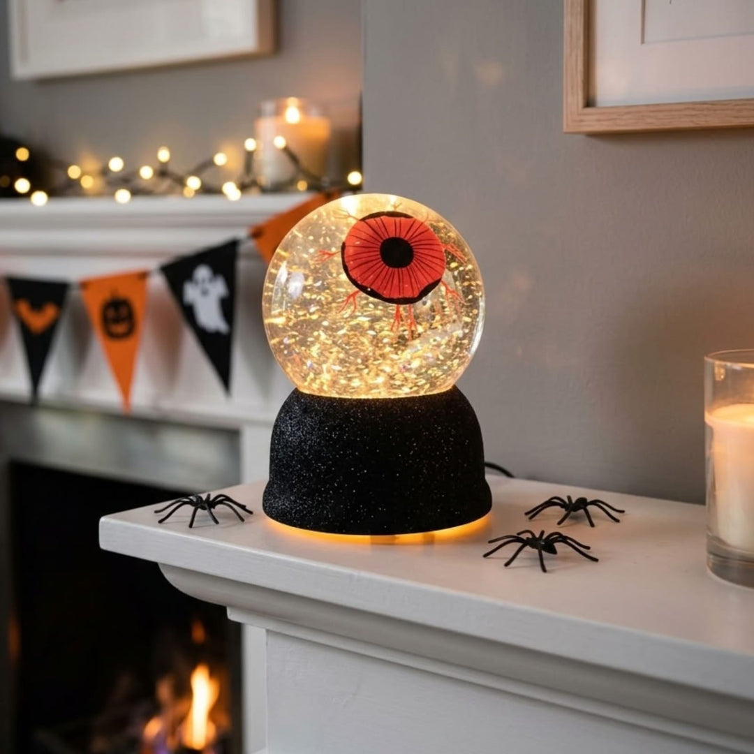 A Halloween-themed decorative snow globe lamp sits on a white fireplace mantel. The clear globe contains glowing warm lights and a large red-and-black eyeball design, mounted on a black glittery base.