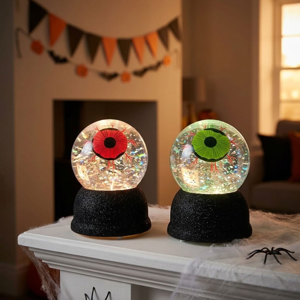 Two Halloween-themed eye snow globes on black bases, displayed on a mantelpiece in a decorated living room. One globe has a red eye and the other has a green eye, both filled with glitter.