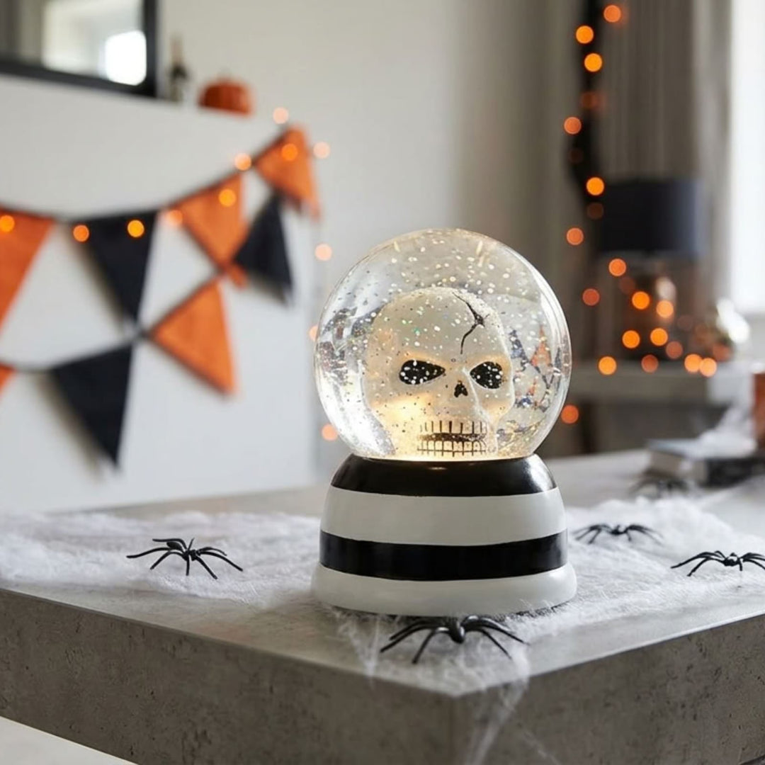 A Halloween-themed snow globe with a skull design inside, sitting on a gray stone surface decorated with fake spiderwebs and black spiders. The background shows a softly blurred indoor setting with orange bokeh lights and a black, white, and orange triangular banner.